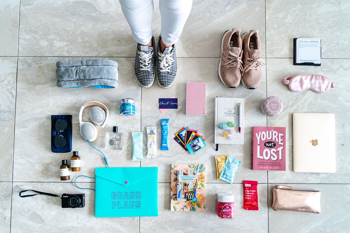 Fashion and travel blogger Jessica Sturdy of Bows & Sequins shares a packing flat lay photo for what she brought along on Remote Year. She's wearing gingham sneakers and standing in the photo with white jeans surrounded by Bose headphones, books, vitamins, RXBars, polaroid photos, and other wellness items that focus on fitness and mindfulness.