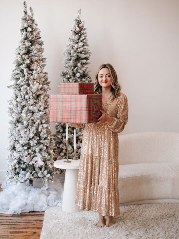 Fashion blogger Jessica Sturdy wearing a gold sequin maxi dress in front of white flocked Christmas trees and a boucle couch holding a stack of presents wrapped in tartan plaid paper.